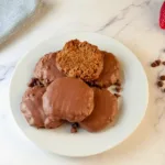 A plate of six keto salted caramel chocolate cookies, with one cookie broken in half to show the soft interior, is placed next to fresh strawberries and chocolate chips on a marble surface.