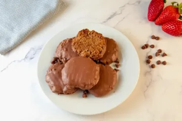 A plate of six keto salted caramel chocolate cookies, with one cookie broken in half to show the soft interior, is placed next to fresh strawberries and chocolate chips on a marble surface.