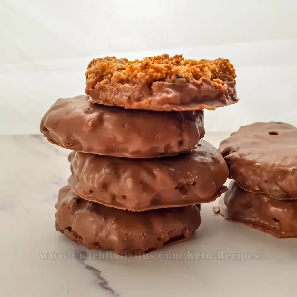 A stack of six keto salted caramel chocolate cookies on a white plate, with caramel sauce drizzled over them and chocolate chips sprinkled on top.