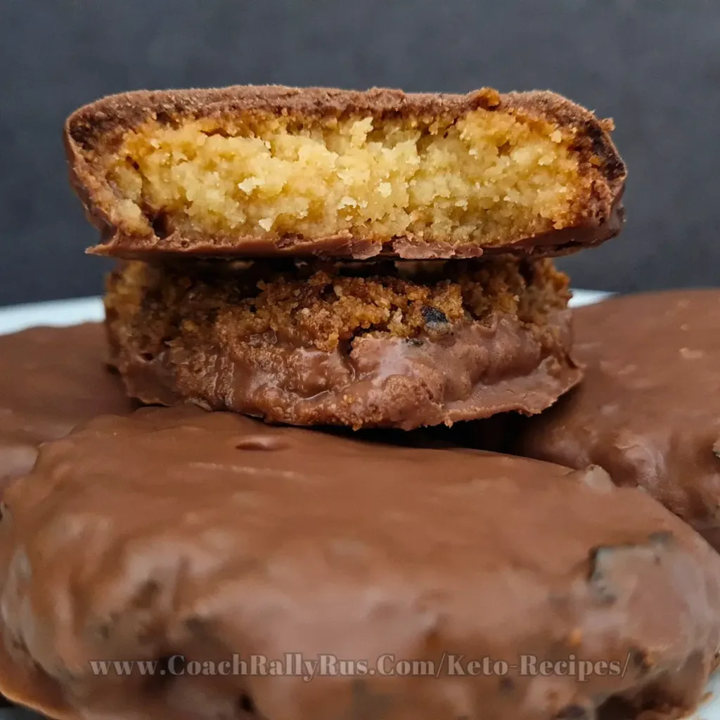 A close-up image of six keto salted caramel chocolate cookies, with one cookie cut in half to show the soft, golden caramel interior, stacked on top of whole cookies that are enrobed in glossy chocolate.