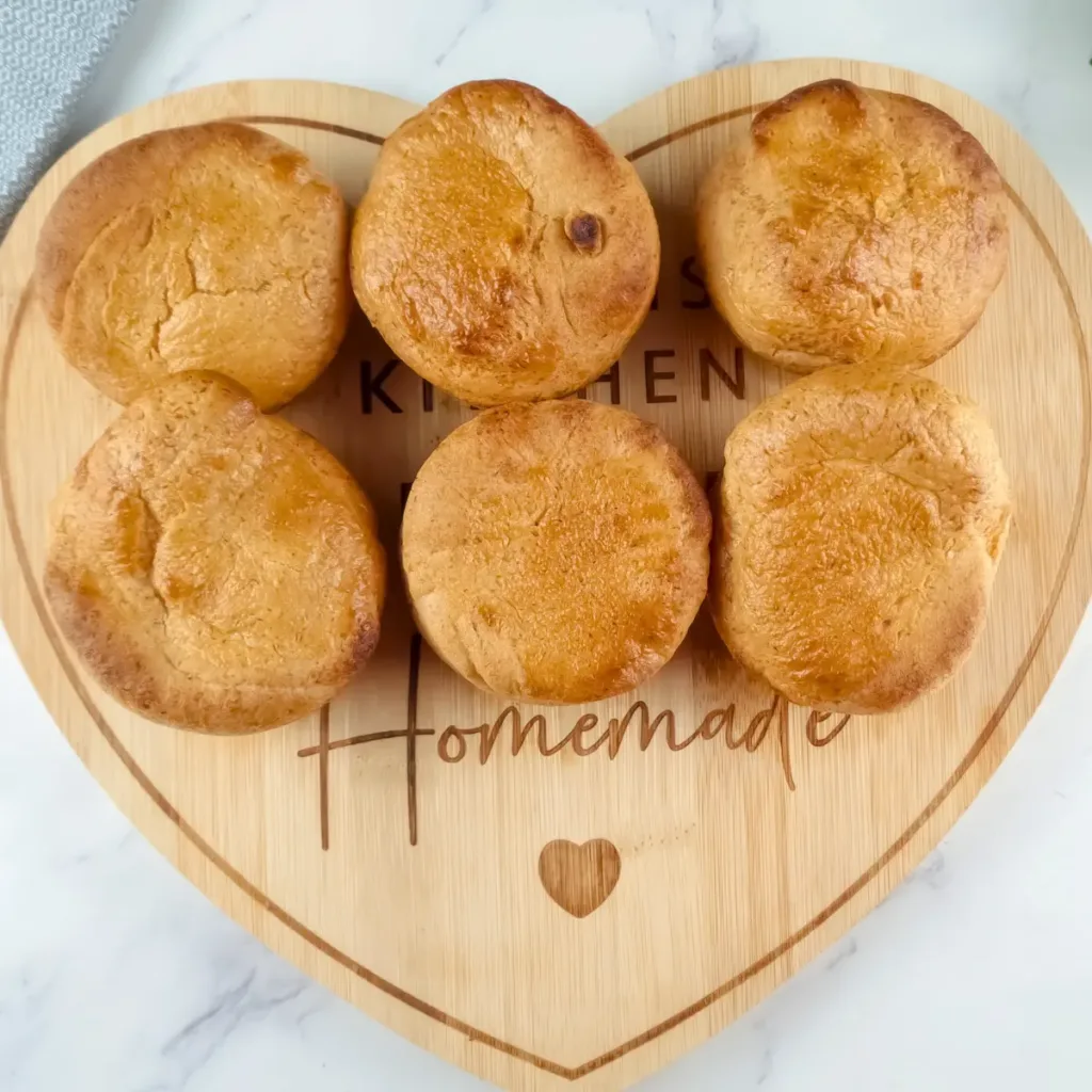 A top view of six golden brown Yorkshire puddings made with almond flour, arranged on a heart-shaped wooden board engraved with the words "KITCHEN Homemade" and a small heart symbol, placed on a white marble surface beside a grey cloth and fresh green parsley.