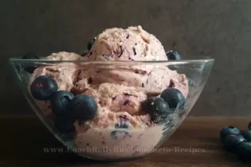 A bowl of high-protein blueberry cottage cheese ice cream, garnished with fresh blueberries on top, presented on a rustic wooden surface with a dark textured backdrop, highlighting the creamy texture and vibrant colors of the dessert.