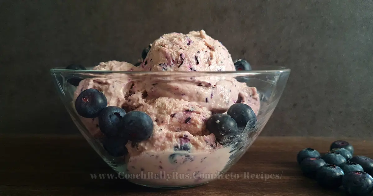 A bowl of high-protein blueberry cottage cheese ice cream, garnished with fresh blueberries on top, presented on a rustic wooden surface with a dark textured backdrop, highlighting the creamy texture and vibrant colors of the dessert.