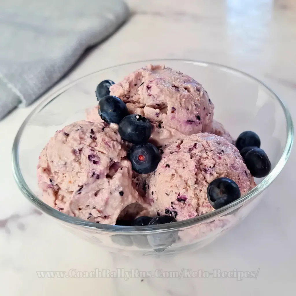 A bowl of high-protein blueberry cottage cheese ice cream, garnished with fresh blueberries on top, presented on a rustic wooden surface with a dark textured backdrop, highlighting the creamy texture and vibrant colors of the dessert.