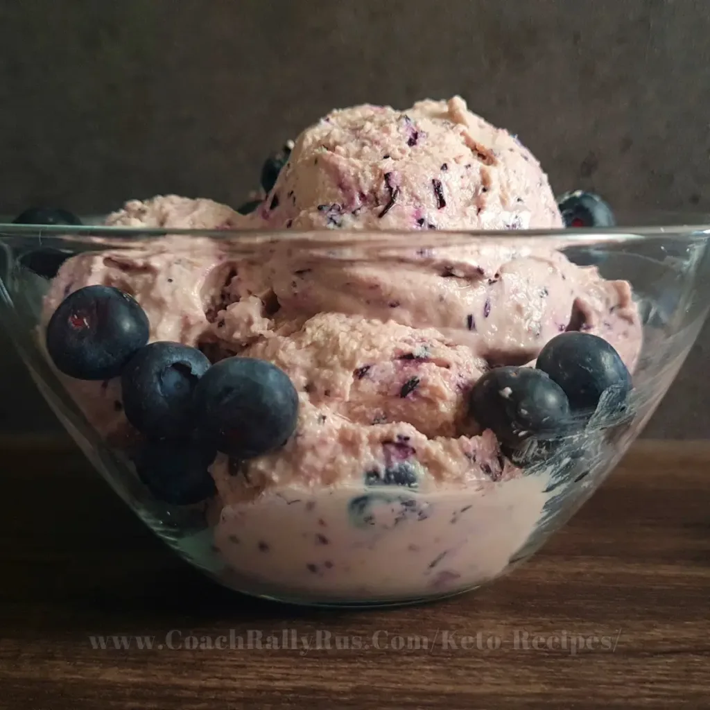 A bowl of high-protein blueberry cottage cheese ice cream, garnished with fresh blueberries on top, presented on a rustic wooden surface with a dark textured backdrop, highlighting the creamy texture and vibrant colors of the dessert.