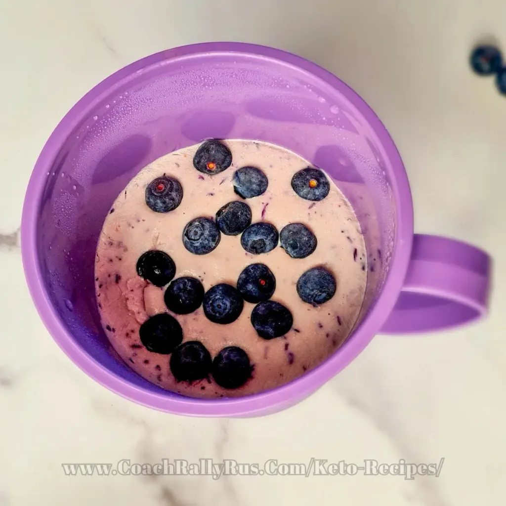 A top view of a purple container filled with blueberry cottage cheese ice cream, placed on a white surface. The image is partially obscured, suggesting a focus on the vibrant purple ice cream with a creamy texture, indicative of its high-protein content.