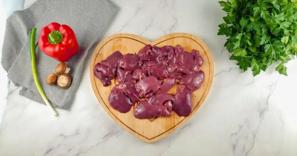 A photo of raw chicken liver arranged on a heart-shaped wooden board, accompanied by fresh vegetables including a red bell pepper, green onion, mushrooms on a grey cloth, and a bunch of parsley on a marble countertop.