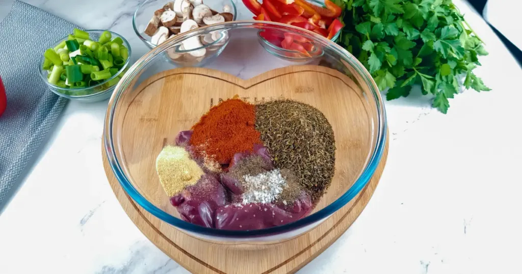 A photo of raw chicken liver arranged on a heart-shaped wooden board, accompanied by fresh vegetables including a red bell pepper, green onion, mushrooms on a grey cloth, and a bunch of parsley on a marble countertop.