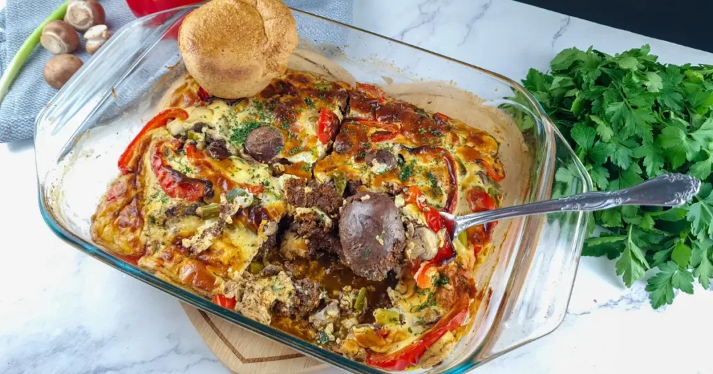 A close-up photo of a baked low carb casserole dish with a golden and creamy topping, filled with chicken liver and vegetables. The keto casserole is garnished with parsley and served on a white plate with a fork. The background is a wooden table with a napkin and a glass of water.
