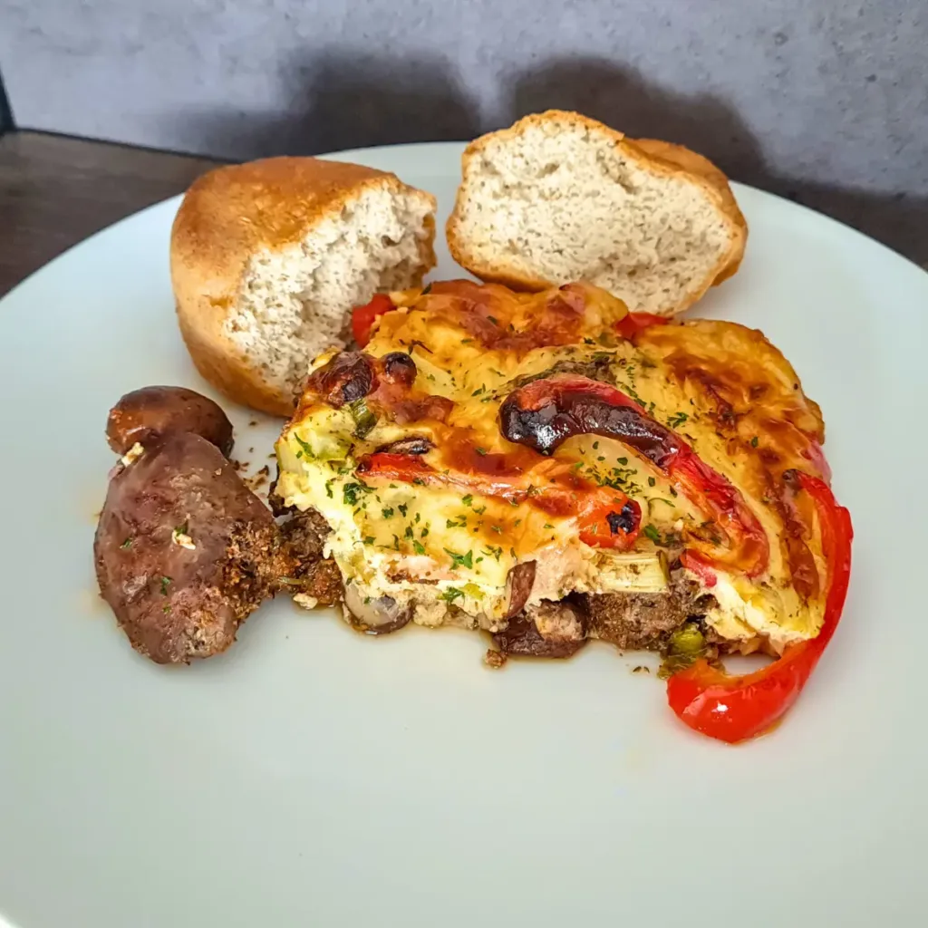 A delicious serving of Keto Chicken Liver Casserole garnished with herbs, accompanied by grilled sausages and freshly baked low carb bread, presented on a white plate.