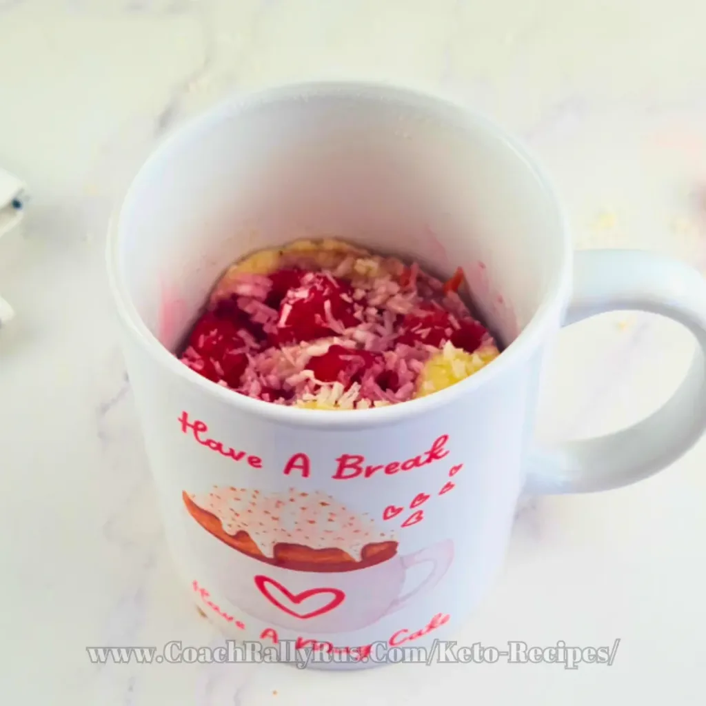 A delicious keto mug cake made with coconut flour and topped with raspberries and shredded coconut, presented on a white plate beside a mug with “Have A Break” text and a donut image, showcasing a tasty yet healthy dessert option.