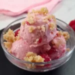 Delicious serving of raspberry cottage cheese ice cream, garnished with crumbled topping, presented in a clear bowl on a black slate, with fresh raspberries and almonds in the background.
