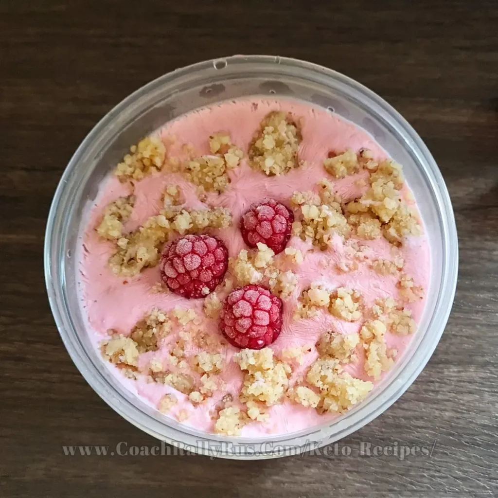 A top view of a container of raspberry cottage cheese ice cream, garnished with whole raspberries and crumbled topping, presented on a wooden surface; the image is labeled with the website www.CoachRallyRus.com/KetoRecipes.