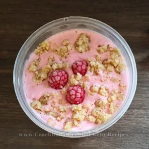 A top view of a container of raspberry cottage cheese ice cream, garnished with whole raspberries and crumbled topping, presented on a wooden surface; the image is labeled with the website www.CoachRallyRus.com/KetoRecipes.