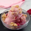 A delicious serving of raspberry cottage cheese ice cream, garnished with crumbled topping, presented in a clear bowl with fresh raspberries and a website link at the bottom