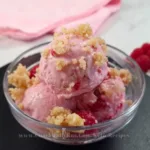 A delicious serving of raspberry cottage cheese ice cream, garnished with crumbled topping, presented in a clear bowl with fresh raspberries and a website link at the bottom