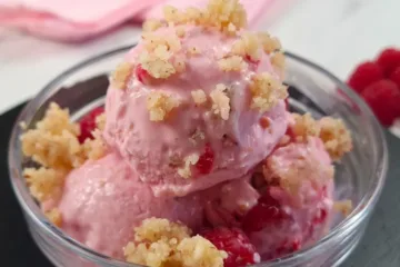 A delicious serving of raspberry cottage cheese ice cream, garnished with crumbled topping, presented in a clear bowl with fresh raspberries and a website link at the bottom