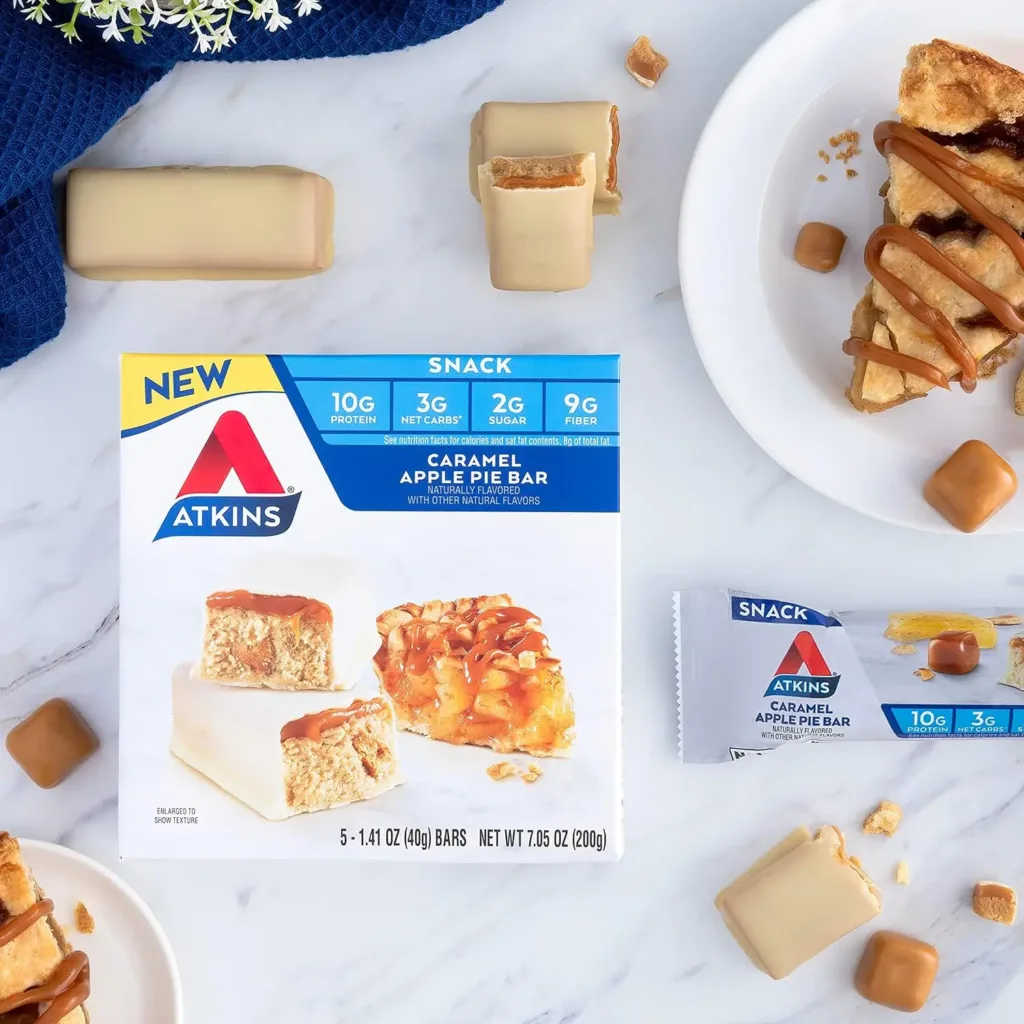 An Atkins Caramel Apple Pie Bar is displayed in its packaging, with pieces of the bar and a slice of apple pie arranged around it on a marble surface, illustrating the snack’s flavor and ingredients.