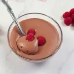 Velvety whipped cottage cheese chocolate mousse topped with a fresh raspberry, accompanied by more raspberries on a sleek marble countertop, showcasing a keto-friendly dessert.