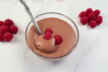 Velvety whipped cottage cheese chocolate mousse topped with a fresh raspberry, accompanied by more raspberries on a sleek marble countertop, showcasing a keto-friendly dessert.