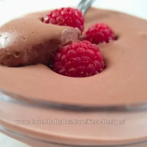 Velvety whipped cottage cheese chocolate mousse topped with a fresh raspberry, accompanied by more raspberries on a sleek marble countertop, showcasing a keto-friendly dessert.