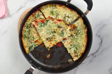 Golden-brown Keto Cottage Cheese Frittata with Pancetta and Broccoli in a black skillet, served on a white marble countertop beside a pink cloth.