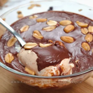 Indulgent Keto Snickers Mousse with layers of smooth cottage cheese and peanut butter blend, topped with a rich chocolate sauce and garnished with whole peanuts, served in a clear glass bowl with a spoonful being lifted, highlighting the dessert’s creamy consistency