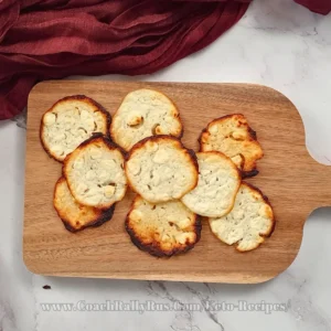 A variety of golden-brown cottage cheese chips with a bubbly surface and crispy edges, displayed on a wooden board against a marble countertop, from CoachRallyRus.com’s keto recipe collection.