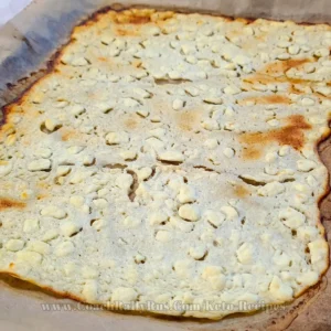 A golden-brown cottage cheese flatbread, freshly baked and full of texture with small, white lumps of cottage cheese visible, sits on parchment paper inside a baking tray, showcasing its homemade, high-protein, and low-carb qualities.