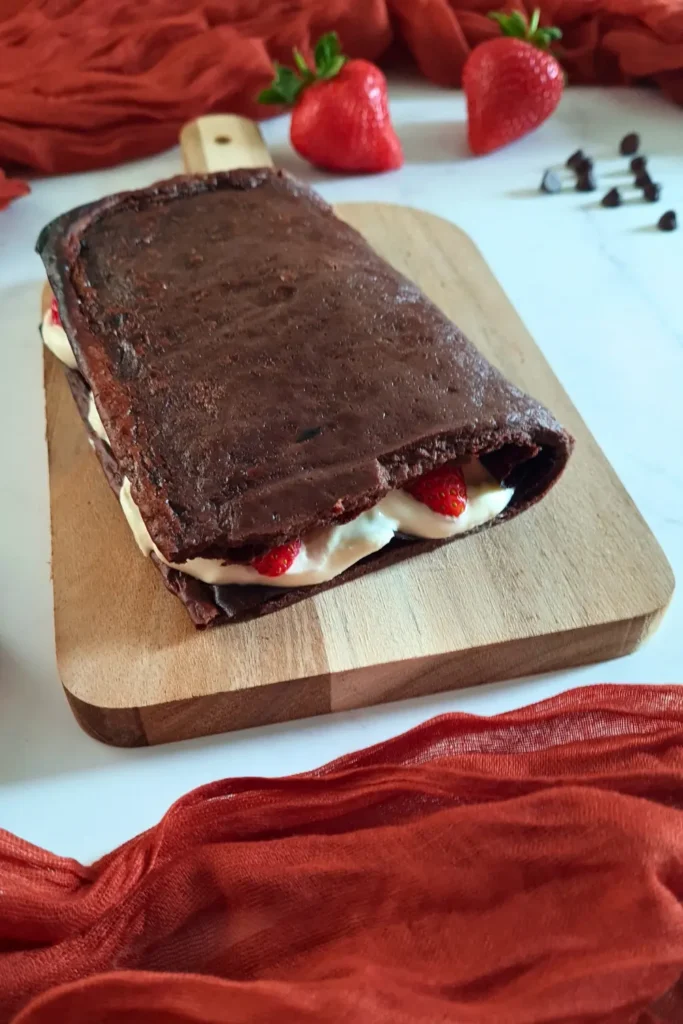 An appetizing chocolate cottage cheese flatbread, featuring rich dark chocolate layers and creamy white cottage cheese, adorned with vibrant red strawberry slices, presented on a wooden cutting board. The composition is enhanced by a scattering of chocolate chips and additional strawberries, set against a backdrop of a ruffled red cloth, creating a visually appealing and indulgent dessert scene.