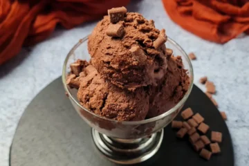 A clear glass bowl filled with scoops of rich Keto Chocolate Ice Cream, presented on a slate coaster. The ice cream has a creamy texture and is garnished with small chocolate chunks. In the background, there’s a soft-focus view of a grey countertop and an orange cloth, which adds a warm contrast to the cool tones of the dessert.