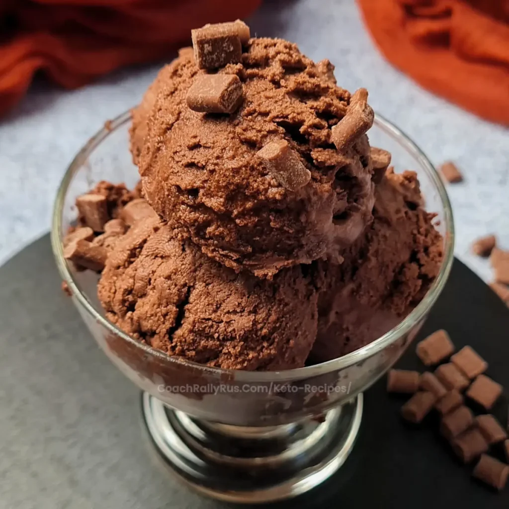 A clear glass bowl filled with scoops of Ninja Creami Keto Chocolate Ice Cream, garnished with chocolate chunks on top. The bowl is placed on a dark grey surface with a rust-colored cloth in the background. In the corner, there’s a watermark that reads ‘coachallyrus.com/keto-recipes’.