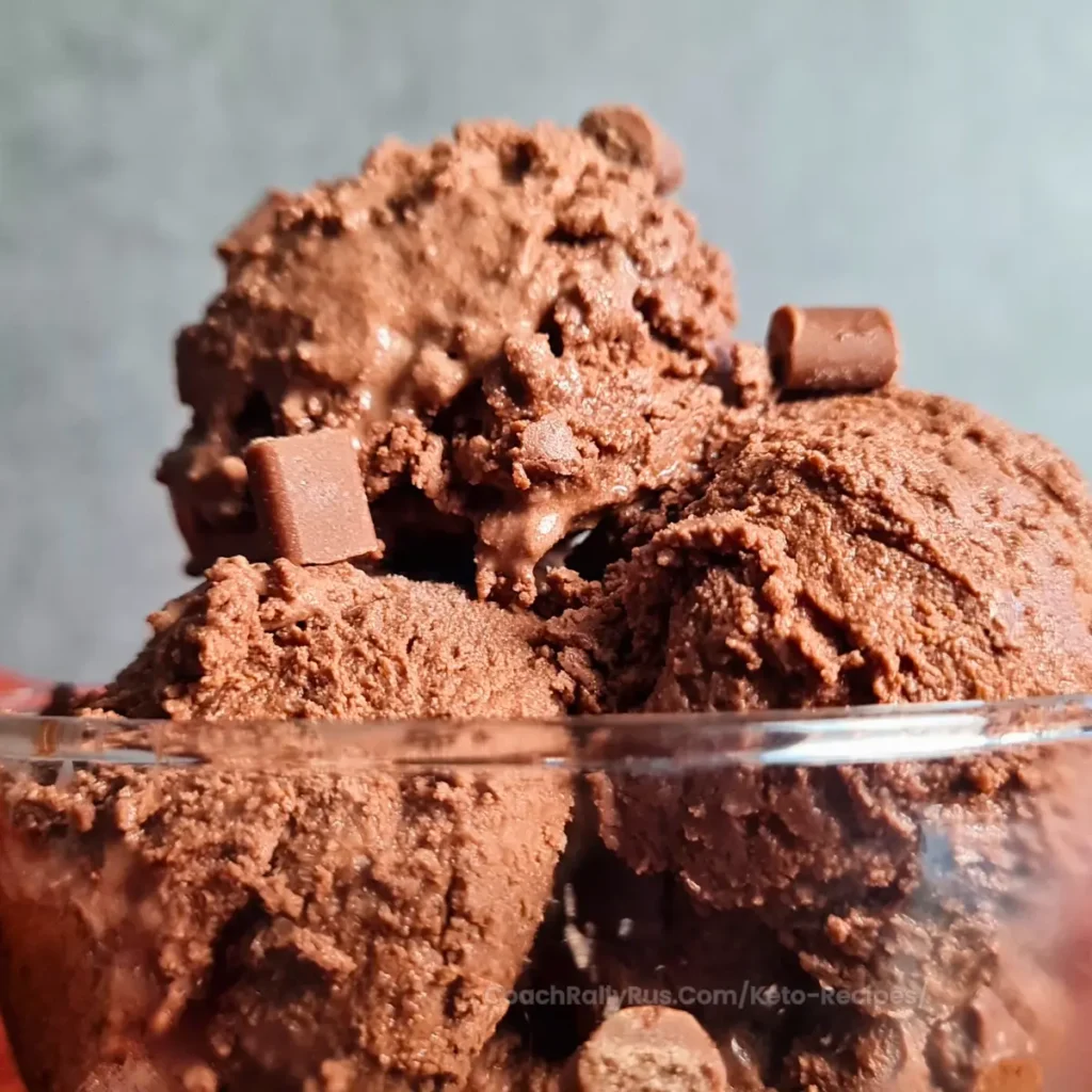Close-up image of Ninja Creami Keto Chocolate Ice Cream served in a clear bowl, showcasing its creamy texture and rich chocolate color. Small chocolate pieces are visible throughout, adding to the indulgent appearance of the keto-friendly dessert. The background is dark, emphasizing the ice cream as the focal point