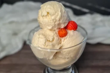 A clear glass bowl filled with two scoops of creamy Ninja Creami Keto Vanilla Ice Cream, garnished with two fresh strawberries on top. The bowl is placed on a dark surface with a white cloth in the background, highlighting the rich texture and pale color of the ice cream, making it an indulgent dessert perfect for a ketogenic diet.
