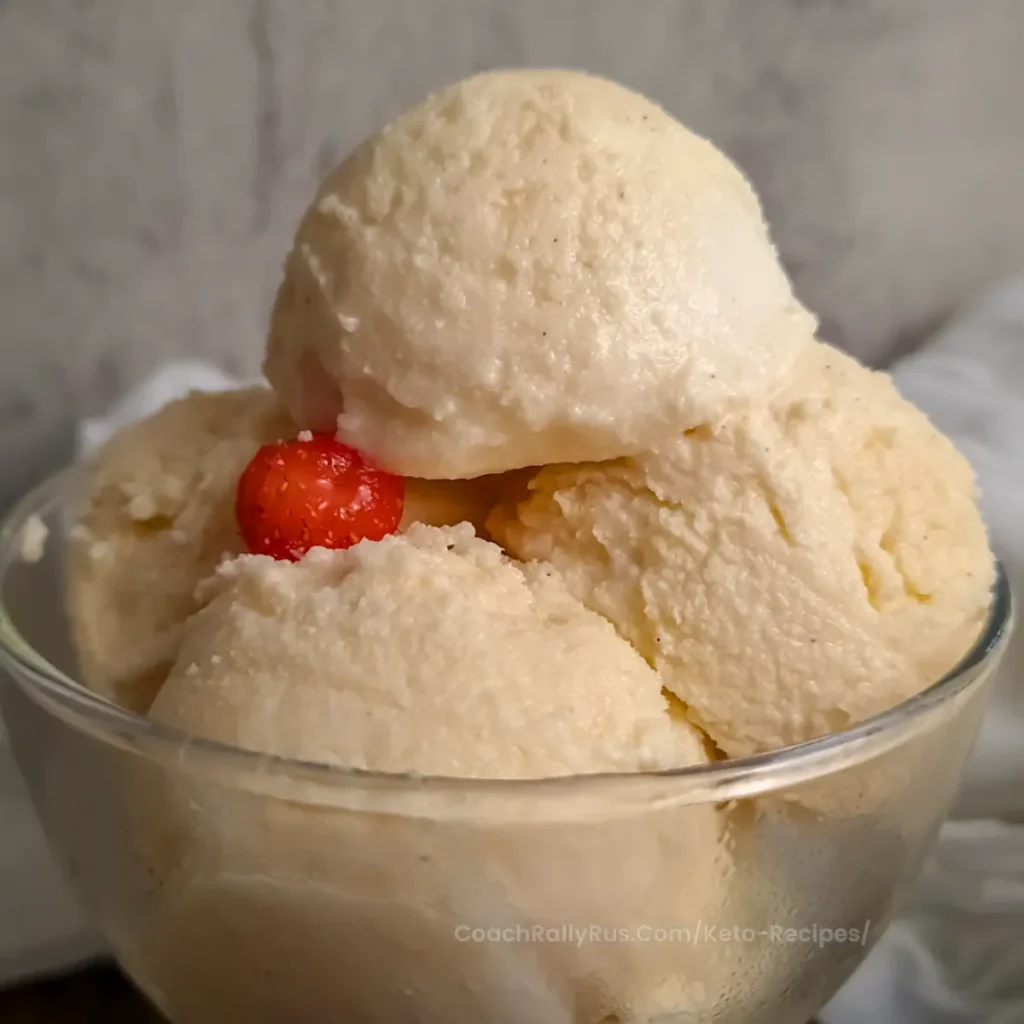 A close-up image of a bowl of Ninja Creami Keto Vanilla Ice Cream, featuring three scoops with a creamy texture. On top of the ice cream, there is a small, ripe red strawberry adding a pop of color. The bowl is set against a soft-focus background that provides a neutral setting to highlight the dessert.