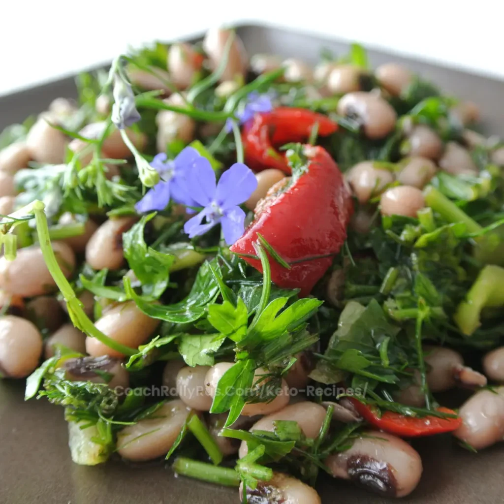 Close-up of a colorful salad featuring black-eyed peas, chopped fresh herbs, and a vibrant purple flower garnish on top. The salad is presented on a dark plate, highlighting the mix of greens, red pepper pieces, and black-eyed peas.
