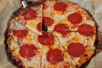 A freshly baked pizza with a golden-brown crust and melted cheese, topped with large pepperoni slices. The pizza is cut into triangular slices and rests on parchment paper with a pizza cutter on top, suggesting it is a homemade or artisanal cottage cheese pizza.