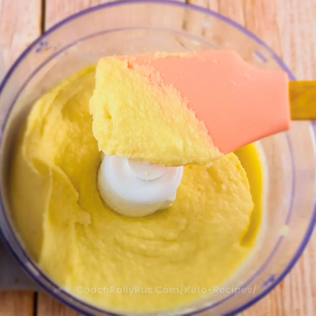 A top-down view of creamy Keto Cauliflower Mash in a food processor with a bright orange spatula. The mash has a smooth, velvety texture and a vibrant yellow color, suggesting the use of butter or cheese for richness. The wooden surface beneath the food processor shows part of the website address ‘CoachRallyRus.Com/Keto-Recipes’.