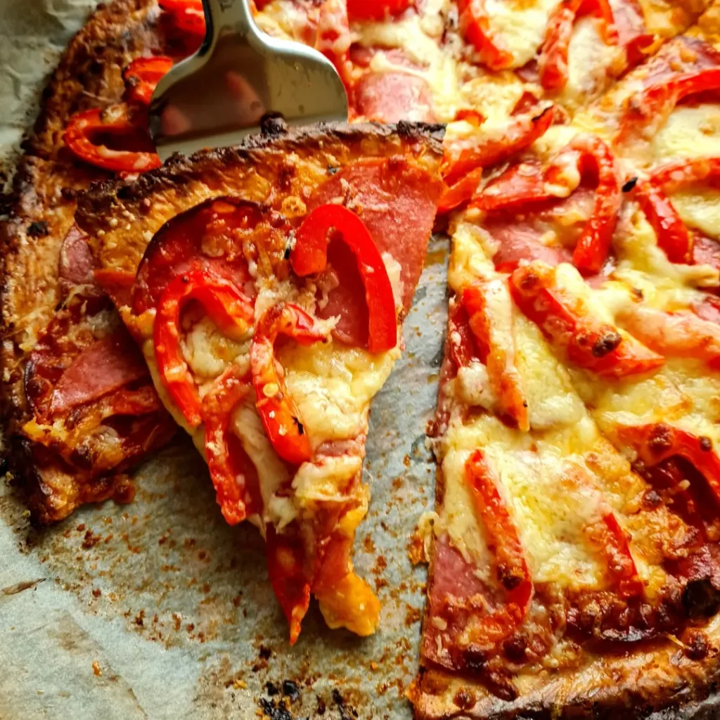 Close-up image of a freshly baked Almond Flour Pizza Crust. The pizza features a golden-brown, keto-friendly fathead dough base, topped with melted cheese, pepperoni slices, and red bell peppers. One slice is being lifted with a metal spatula, showing the stringy cheese and crispy crust. This image highlights a gluten-free, low-carb pizza option perfect for those following a ketogenic diet.