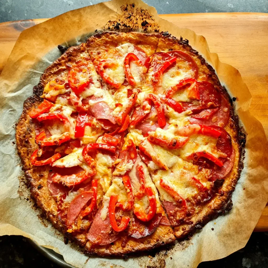 Close-up image of a freshly baked Almond Flour Pizza Crust. The pizza features a golden-brown, keto-friendly fathead dough base, topped with melted cheese, pepperoni slices, and red bell peppers. One slice is being lifted with a metal spatula, showing the stringy cheese and crispy crust. This image highlights a gluten-free, low-carb pizza option perfect for those following a ketogenic diet.