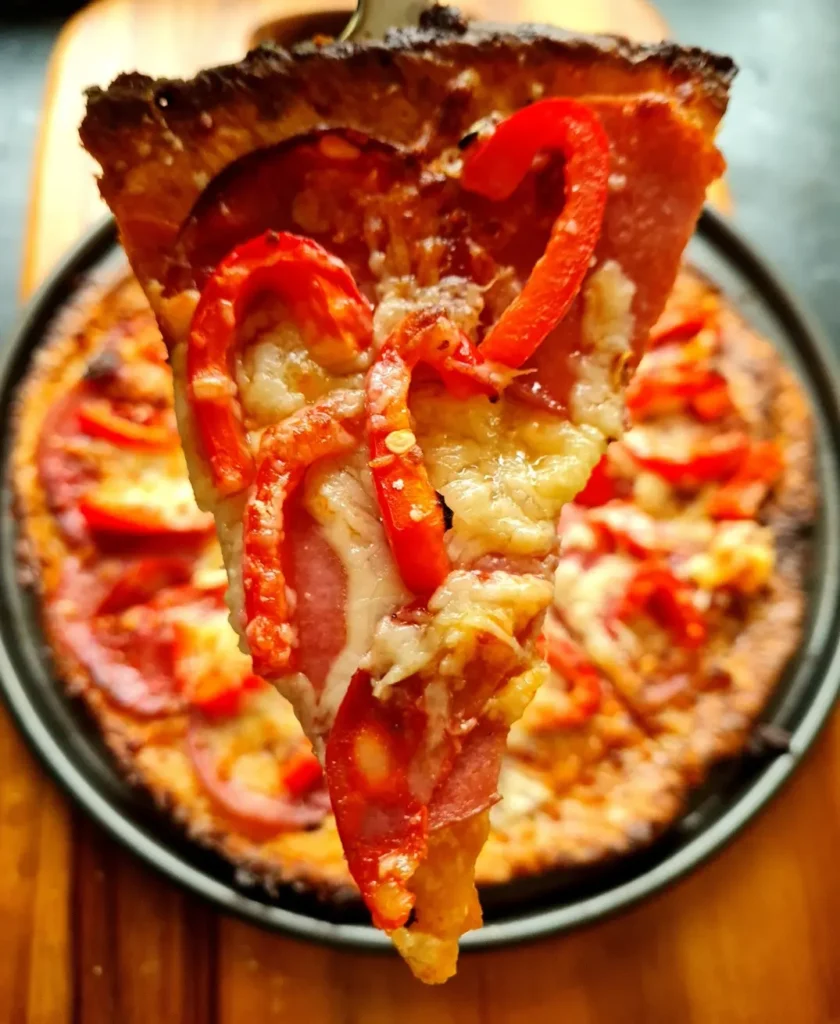 Close-up image of a freshly baked Almond Flour Pizza Crust. The pizza features a golden-brown, keto-friendly fathead dough base, topped with melted cheese, pepperoni slices, and red bell peppers. One slice is being lifted with a metal spatula, showing the stringy cheese and crispy crust. This image highlights a gluten-free, low-carb pizza option perfect for those following a ketogenic diet.