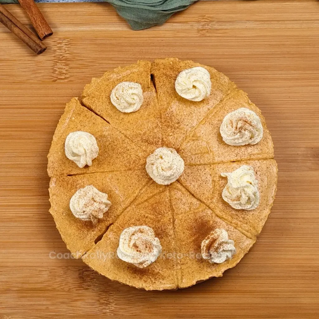 A top view of a Cottage Cheese Pumpkin Cheesecake on a wooden surface, garnished with dollops of whipped cream and cinnamon sticks. The cheesecake is marked into even slices, ready to be served.