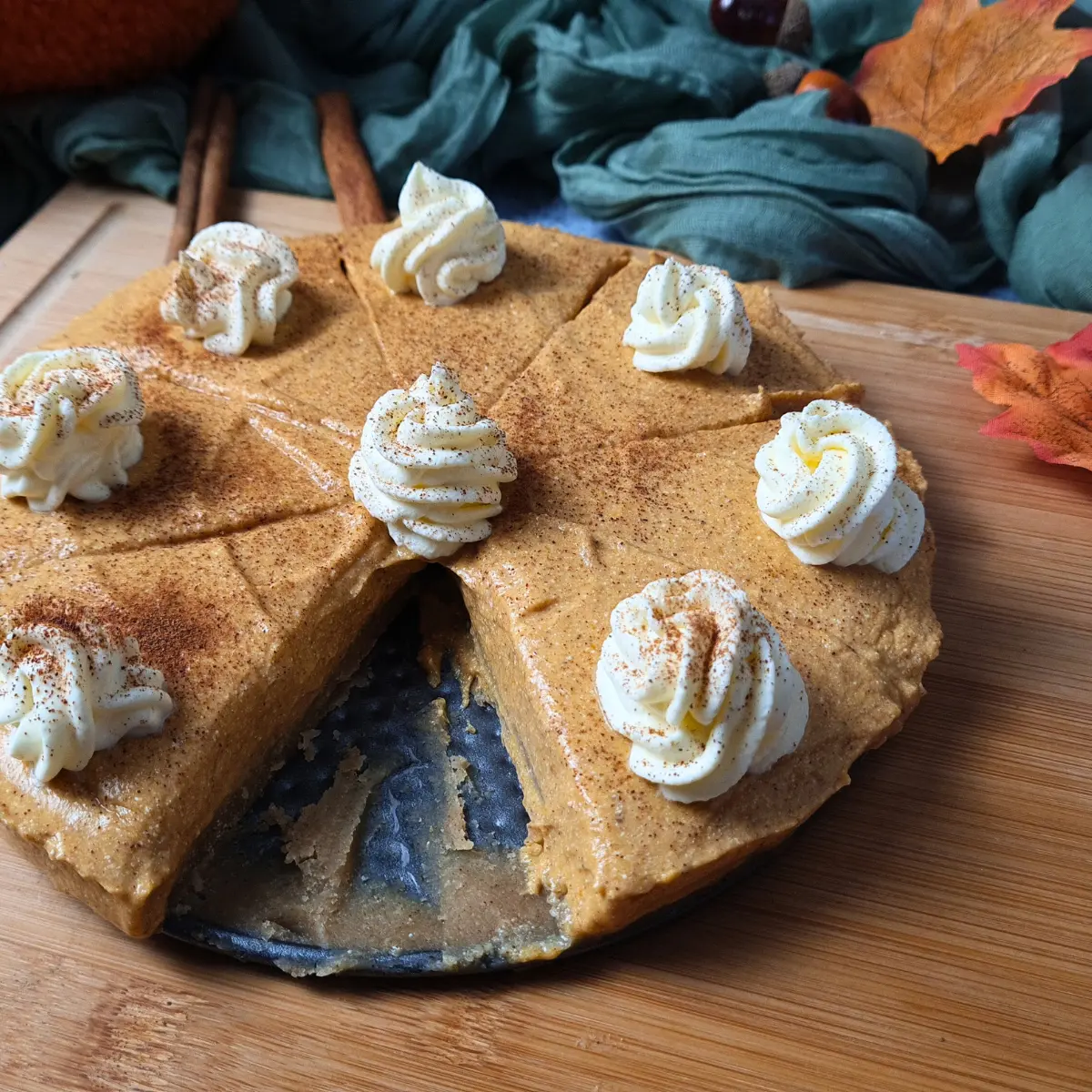 A Cottage Cheese Pumpkin Cheesecake on a wooden surface, with one slice removed, topped with whipped cream and garnished with cinnamon sticks.