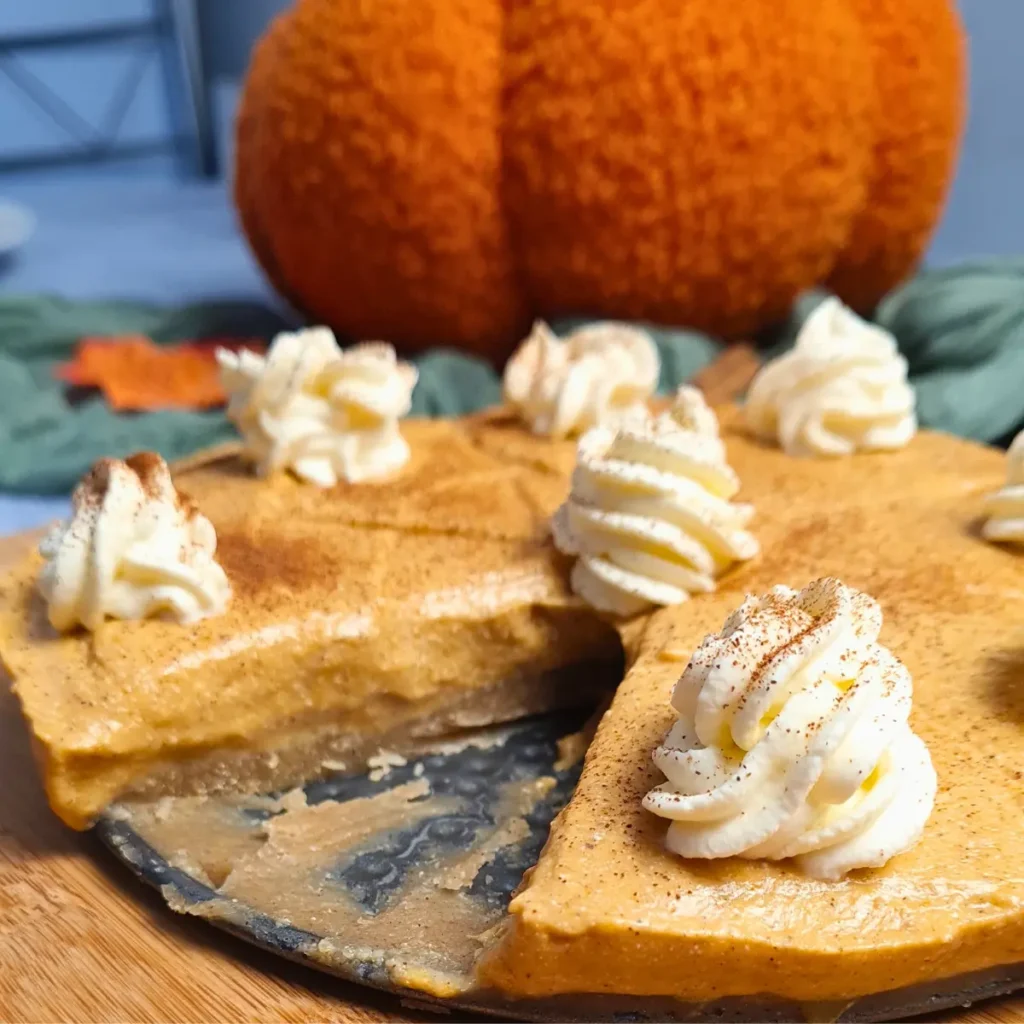 A close-up image of a Cottage Cheese Pumpkin Cheesecake with one slice partially removed, topped with whipped cream and a dusting of spices.