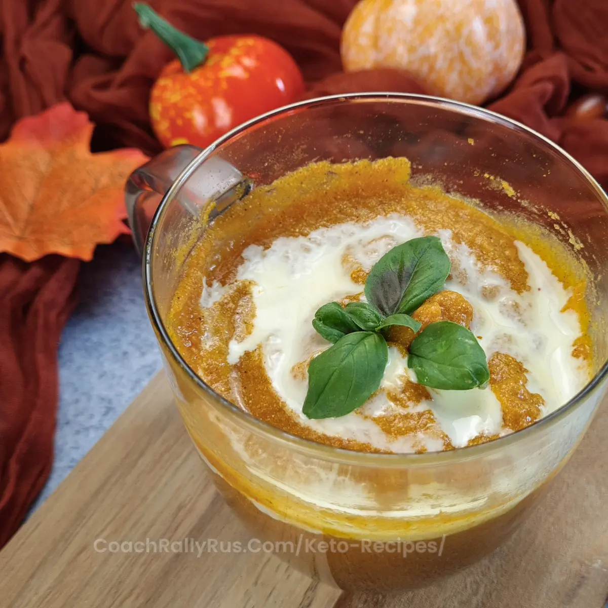 A clear glass bowl of pumpkin soup with a swirl of cottage cheese and a basil leaf garnish, set against a backdrop of autumn leaves and a small pumpkin.