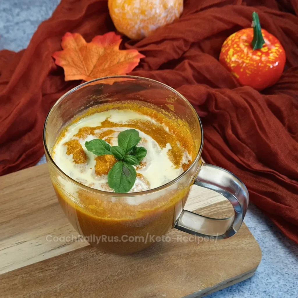 A clear glass mug filled with creamy orange-colored pumpkin soup, garnished with a swirl of cream, a sprinkle of spices, and a fresh basil leaf on top. The mug is placed on a wooden board against a backdrop of a blue textured surface with an autumnal theme including an orange cloth, decorative pumpkins, and fallen leaves.