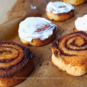 A close-up image of freshly baked keto cinnamon rolls made with coconut flour, topped with a creamy white frosting. One roll is partially unraveled, revealing its spiral layers, and another has a bite taken out of it to show the texture inside. The rolls are placed on a dark surface, and there’s a website address at the bottom: CoachRallyRus.Com/KetoRecipes.