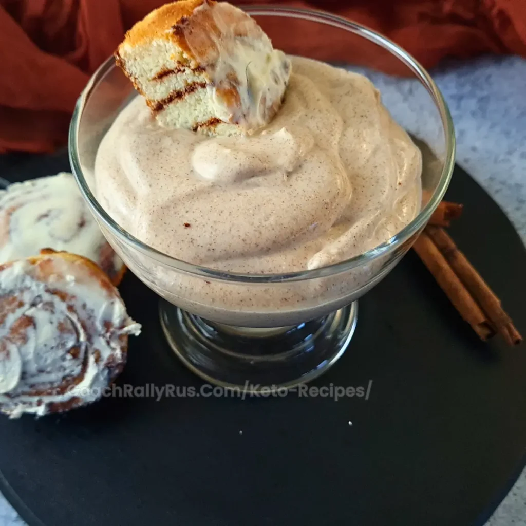 A glass bowl filled with a creamy cinnamon-flavored parfait topped with a piece of cinnamon roll, placed on a black surface. Two cinnamon sticks and additional slices of cinnamon roll are visible beside the bowl.