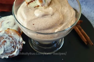 A glass bowl filled with a creamy cinnamon-flavored parfait topped with a piece of cinnamon roll, placed on a black surface. Two cinnamon sticks and additional slices of cinnamon roll are visible beside the bowl.
