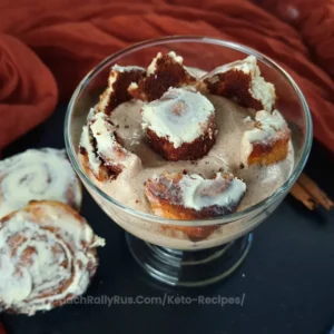 A glass parfait filled with layers of cottage cheese and cinnamon roll pieces, topped with a creamy frosting. The dessert is presented on a dark red cloth background, with a whole cinnamon roll and sticks of cinnamon nearby for garnish.